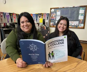 Adult and student sitting together at classroom table holding book 